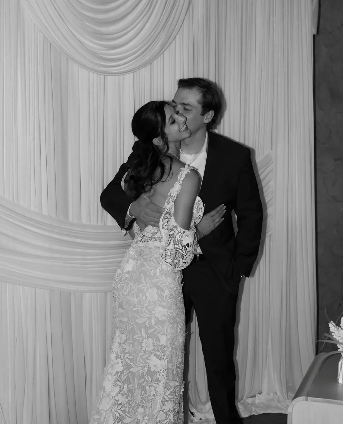 Black and white photo of a groom kissing his bride's cheek in front of ivory drapery