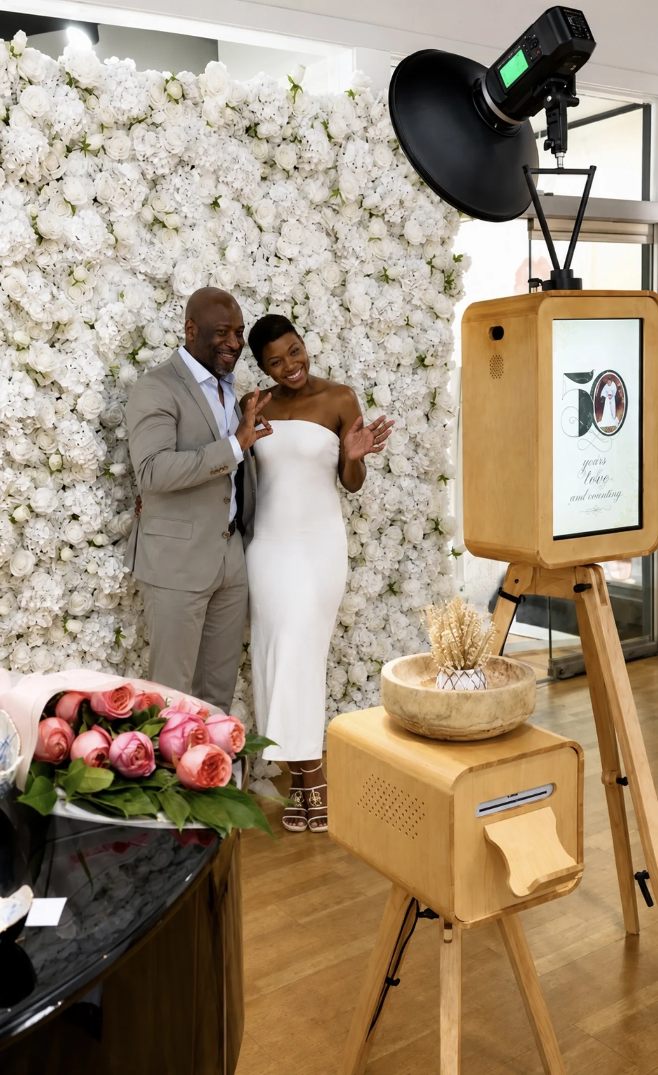 Guests posing at the white flower wall with the Luxury Memories wooden DSLR booth, studio light and printer