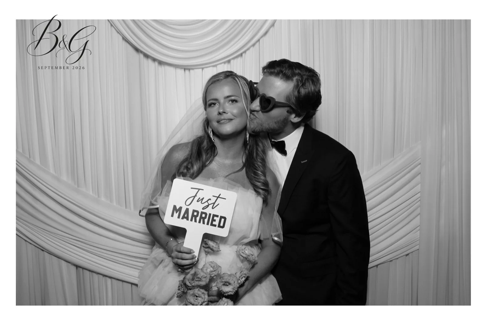Black and white glam portrait: groom in heart sunglasses kisses the bride, who holds a Just Married sign