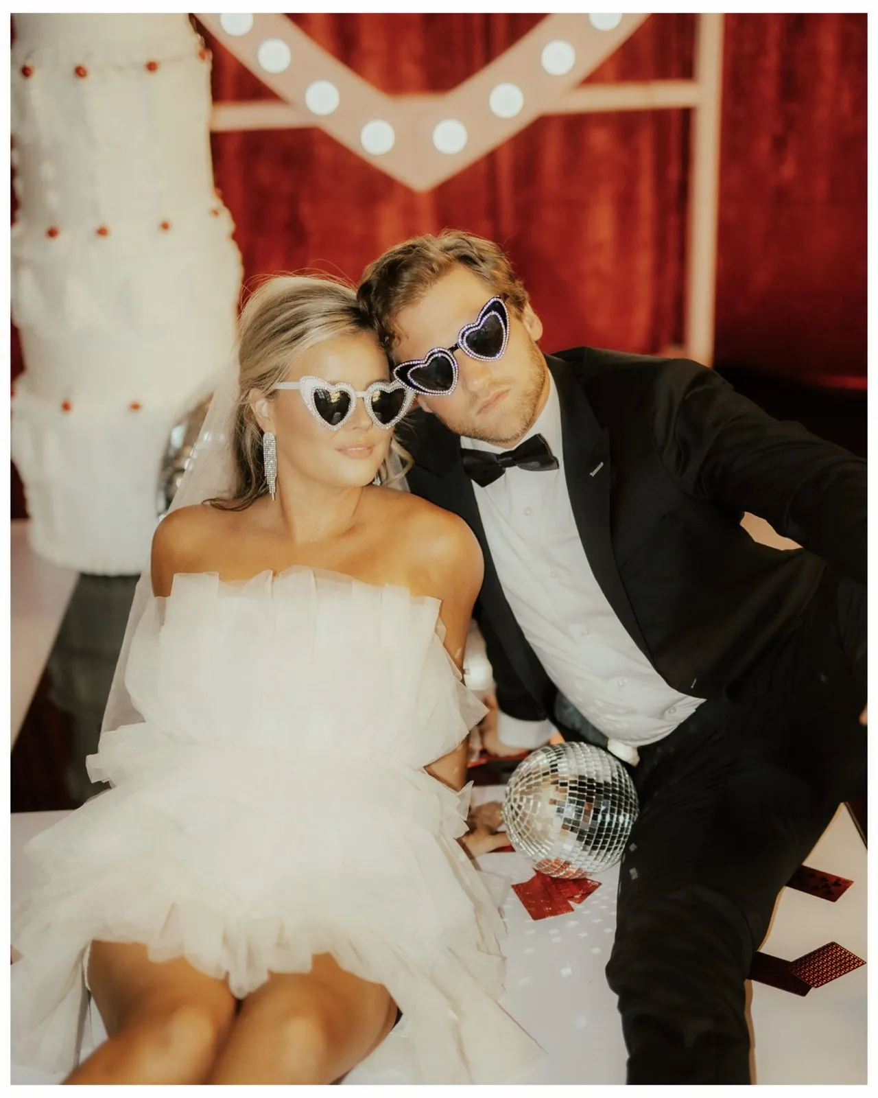 Newlyweds in heart sunglasses in front of red velvet and a marquee heart
